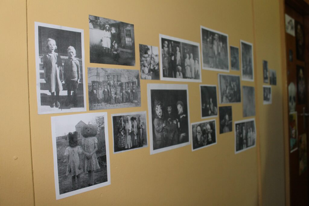 Image shows a sandy coloured wall covered in randomly spaced photogtaphs. The photographs are printouts of creepy vintage black and white halloween photos, sourced from the internet.