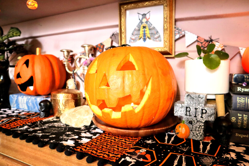 Image shows a Halloween display using sustainable halloween decorations, on top of a vintage halloween table runner. There is a carved jack o'lantern, a papier mache jack o'lantern, a silver candlestick, gravestone shaped candle, small skull shaped candles, indoor plants and vintage books. On the wall behind these objects hangs mini triangle bunting and an original illustration of a death's head hawkmoth in a vintage gold square frame.