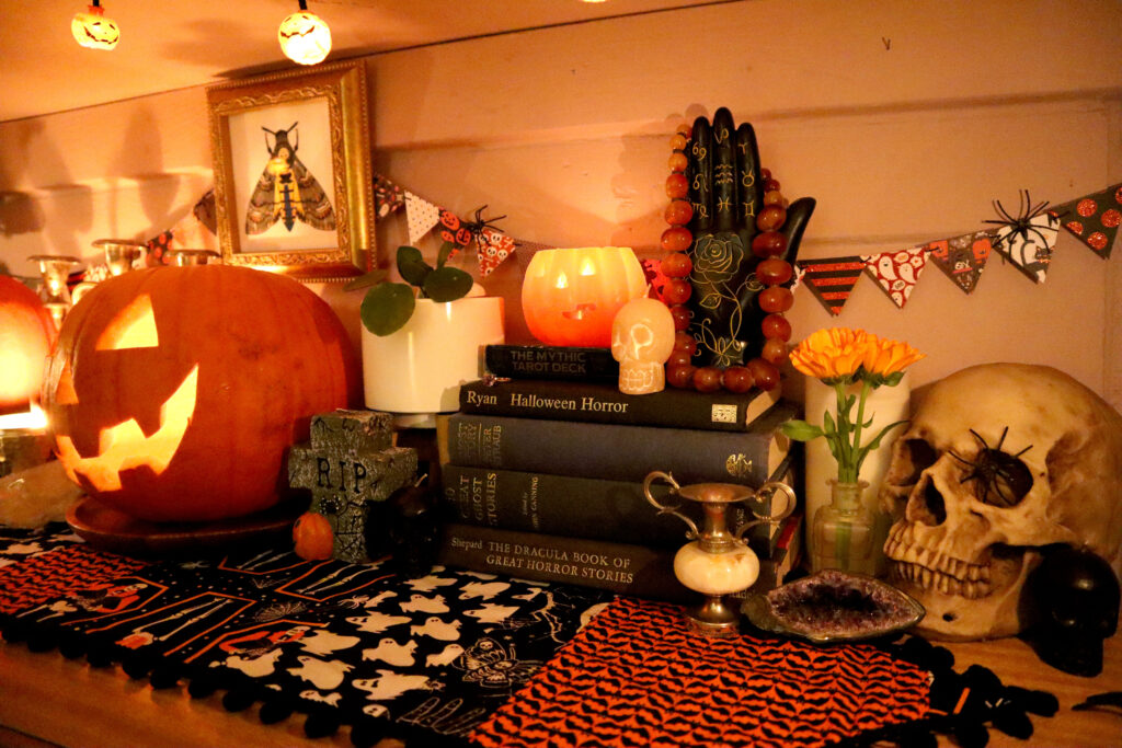 Image shows a Halloween display on top of a vintage halloween table runner. There is a carved jack o'lantern, a gravestone shaped candle, small skull shaped candles, indoor plants, a black palmistry hand, a resin skull, vintage vases, candlesticks, a raw amethyst and a stack of vintage books. On the wall behind these objects hangs mini triangle bunting and an original illustration of a death's head hawkmoth in a vintage gold square frame. The whole scene is dimly lit with a jack o'lantern, candles, and a string of jack o'lantern fairy lights.