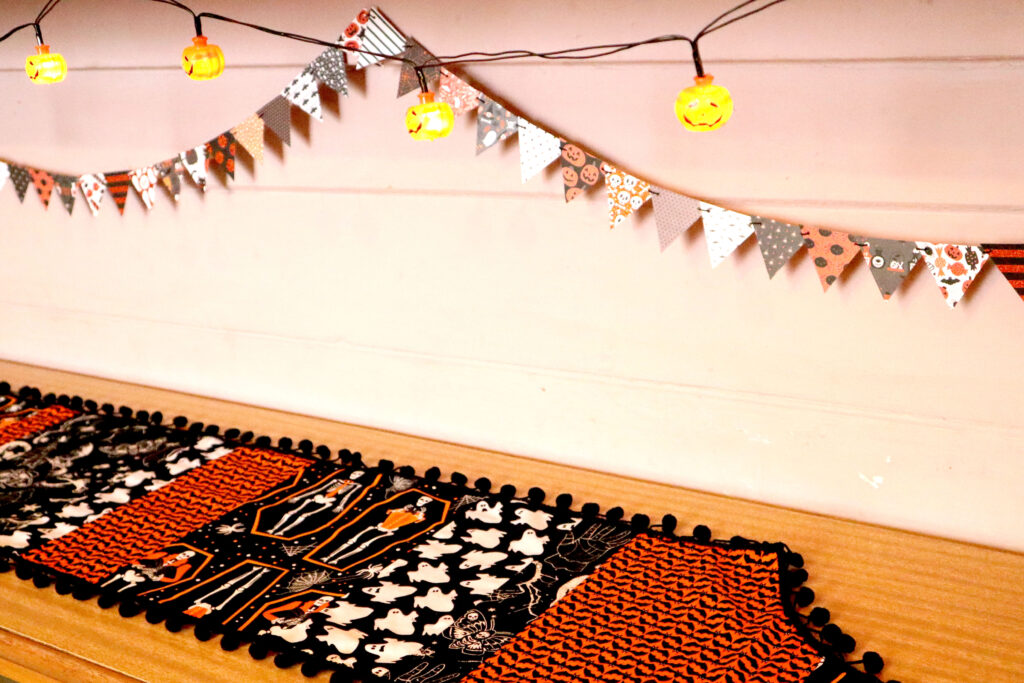 Image shows the base layer of styling a Halloween display, without all the objects. At the back of the display area hangs a string of Halloween Mini Bunting and a string of jack o'lantern fairy lights against a pale pink wall. In front of the wall is a vintage 60s laminate look buffet. The buffet is topped with a vintage halloween table runner.