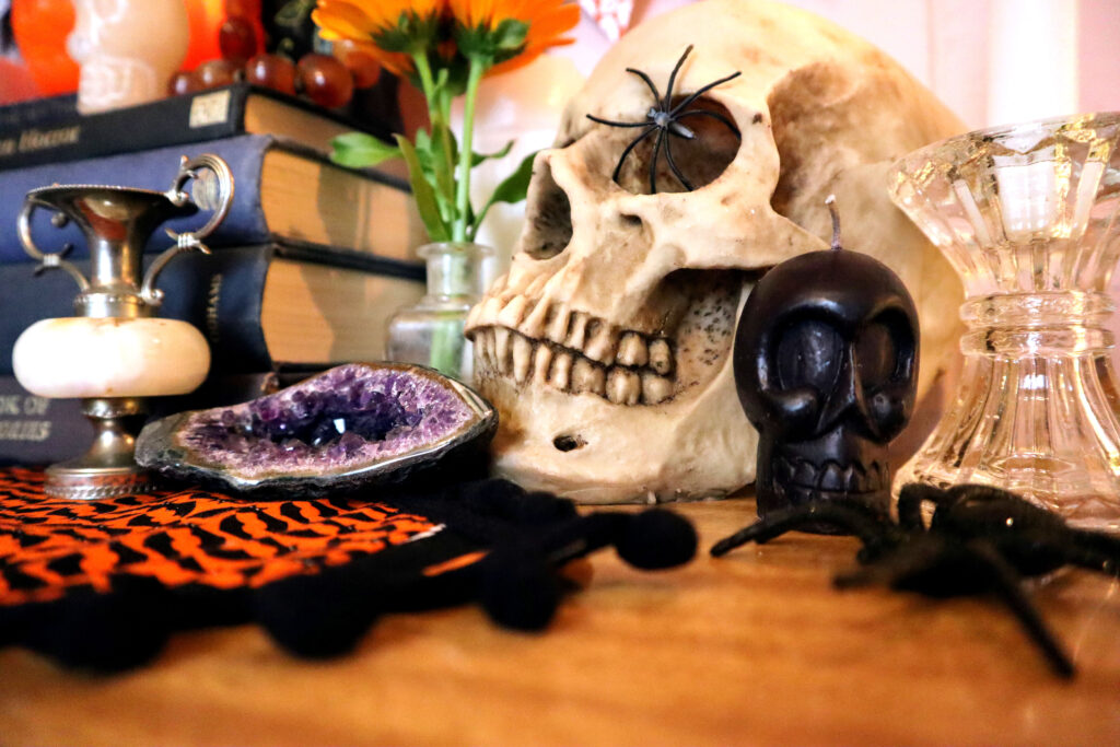 Image shows a halloween display. There is a stack of fabric covered vintage books in indigo shades. To the right of the books is a vintage glass inkwell containing vivid orange calendula flowers. Behind the vase is a white pilar candle. In front of the vase is a raw amethyst crystal and small vintage bud vase. Next to the inkwell, is a realistic resin skull with a black plastic spider in its eye socket. In front of the skull is a small black skull candle, and a glass candlestick holding a tall white candle. A Halloween Table runner is also visible, it sits on a timber-look laminate surface.