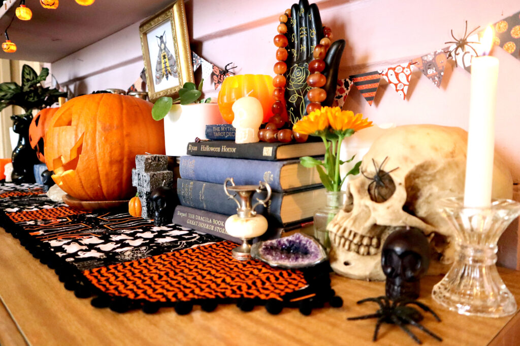 Image shows a Halloween display on top of a vintage halloween table runner. There is a carved jack o'lantern, a papier mache jack o'lantern, a gravestone shaped candle, small skull shaped candles, indoor plants, a black palmistry hand, a resin skull, vintage vases, candlesticks, a raw amethyst and a stack of vintage books. On the wall behind these objects hangs mini triangle bunting and an original illustration of a death's head hawkmoth in a vintage gold square frame.