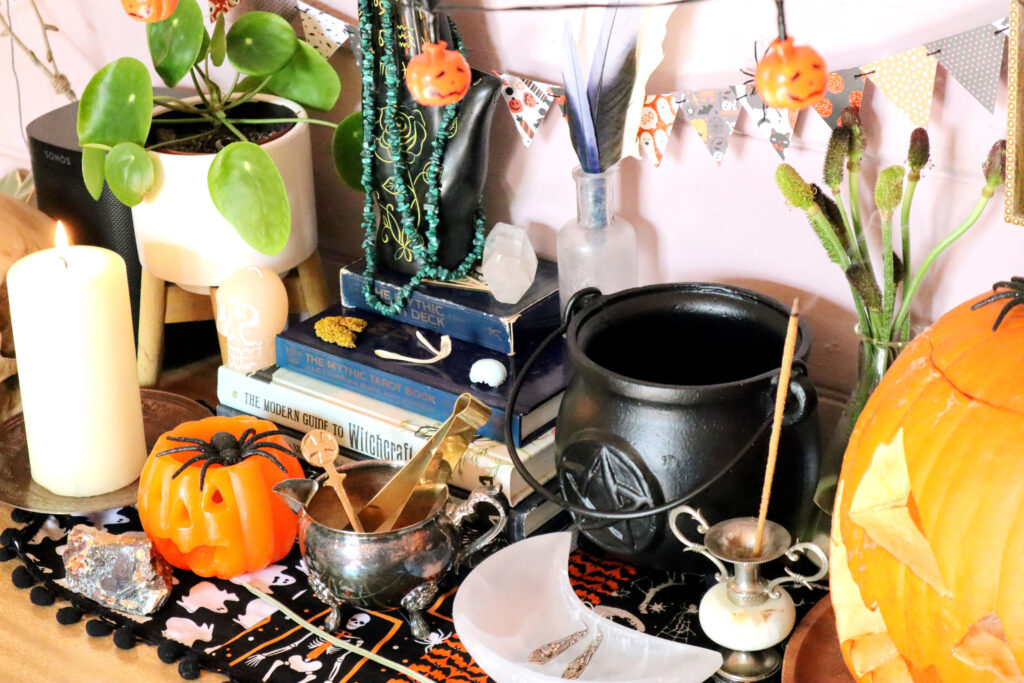 Image shows detail from a Halloween display. In the centre of the image is a stack of witchy books and a box of tarot cards. On top of the stack is a black palmistry hand draped in a green malachite chip necklace. Near the hand is a raw clear quartz crystal, a mustard coloured piece of lichen, a wishbone and a piece of blue eggshell. To the left of the stack of books is a chinese money plant in a small white pot, a bone coloured skull candle, and a large pilar candle on a copper coloured metal dish. In front of the stack of books is an orange jack o'lantern tea holder, a piece of raw orange sphalerite crystal, and a tarnished silver vintage milk jug containing a pair of small brass tongs and a small brass spoon. To the right of the stack is a vintage bottle containing blue, black, yellow and white bird feathers, a black cauldron, a vinatage glass flask containing the stems and seed heads of ranunculus flowers, and a carved jack o'lantern.In front of the cauldron is a vintage metal bud vase and a moon shaped selenite dish containing a pair of silver earrings. All items are sitting on a vintage halloween table runner.