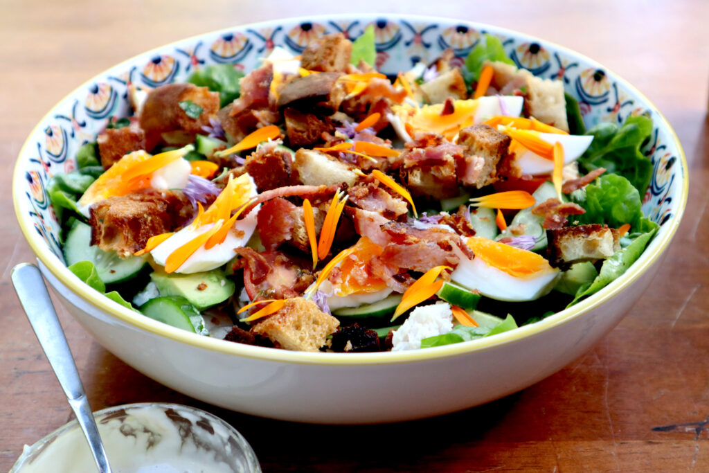 Image show a large salad bowl with a colourful pattern. Inside the salad bowl is a Ceasar Salad. On a bed of cos lettuce, ther are cherry tomatoes, sliced cucumber, sliced avocado, hard boiled eggs cut into wedges, pieces of grilled chicken, and shards of crispy bacon. The salad is garnished with vibrant orange calendula and purple chive petals.