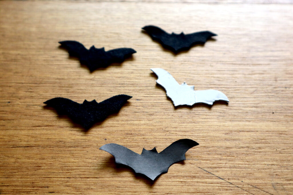 Image shows cut out bat templates and felt bats on a vintage timber desk. There is a paper cutout from Maddy the Maker's free bat template printable, a bat template cut out of recycled white cardboard, and three black felt bats.