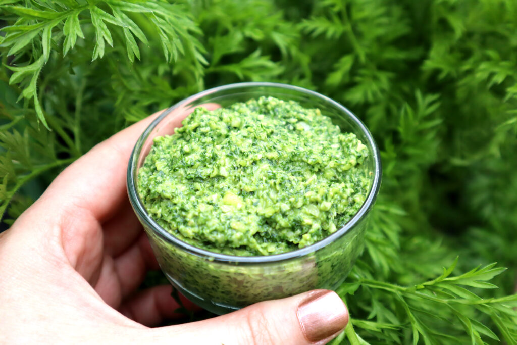 Image shows Maddy the Maker's hand holding a clear glass dish of vibrant green pesto. Maddy is wearing shiny copper coloured nailpolish. The background of the image shows fresh green carrot tops.