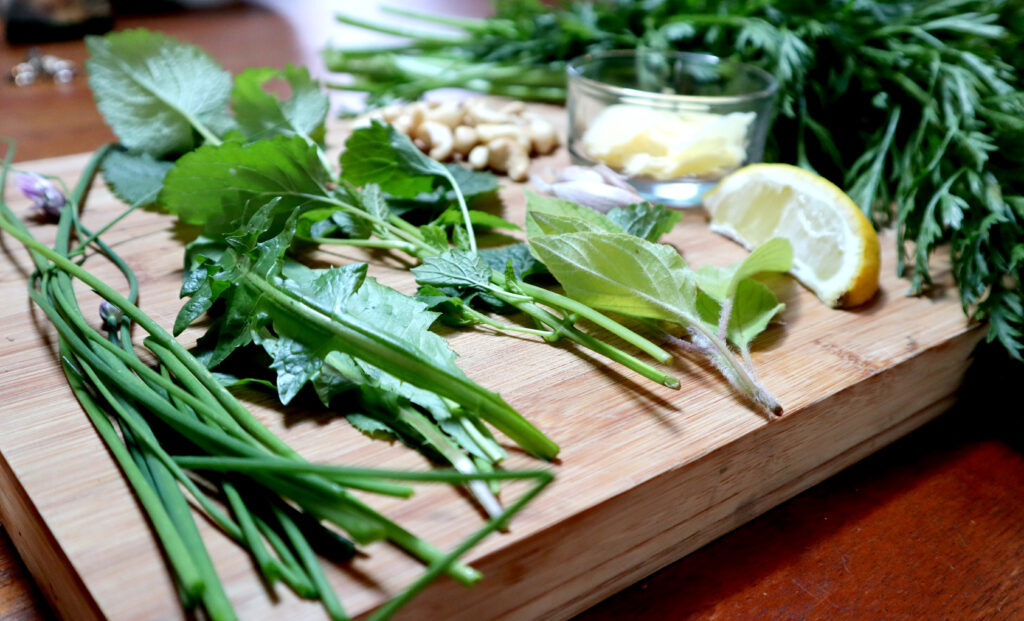 Image shows a bamboo chopping board covered in fresh herbs and other garden ingredients.There are chives, leaves from dandelion, lemon balm, pineapple sage, and carrot tops. There is also a wedge of lemon, a couple of garlic cloves, and a handful of cashew nuts.