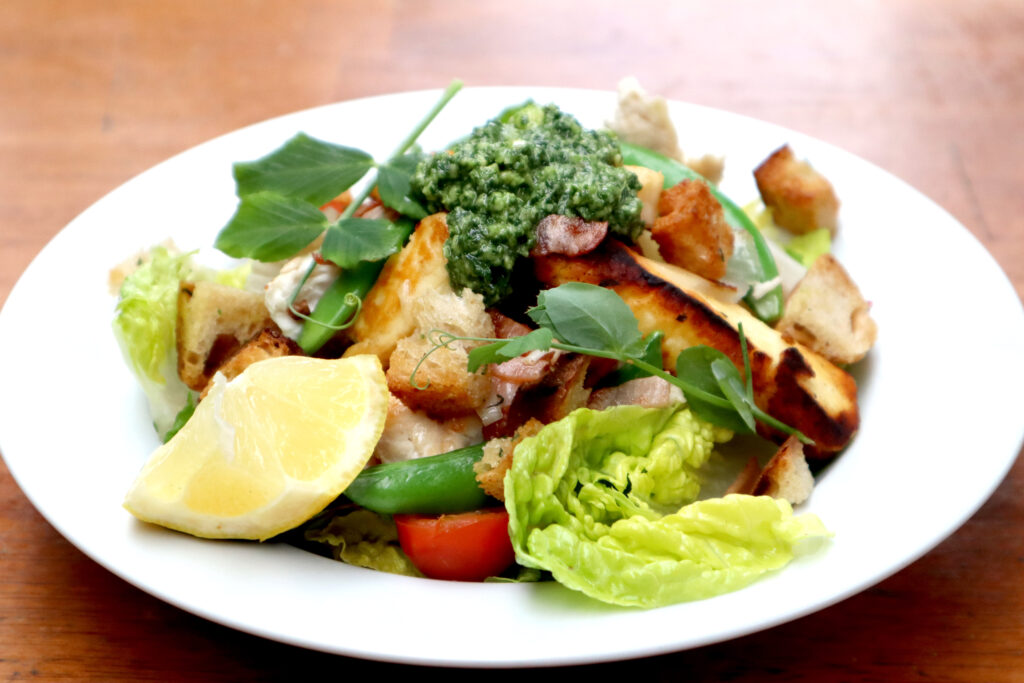 Chicken and Haloumi salad is shown in a white ceramic bowl. The bowl is shown on a timber surface. The salad contains cos lettuce, cherry tomatoes, green beans, fried chicken,  haloumi and bacon, sliced red onion and crunchy herbed croutons. The salad is garnished with a dollop of fresh garden pesto, pea shoots, and a lemon wedge.