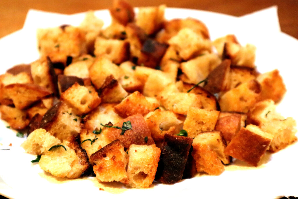 Closeup of Crunchy Herbed Croutons on a white plate. They are crispy golden cubes of fried bread, sprinkled with green herbs.