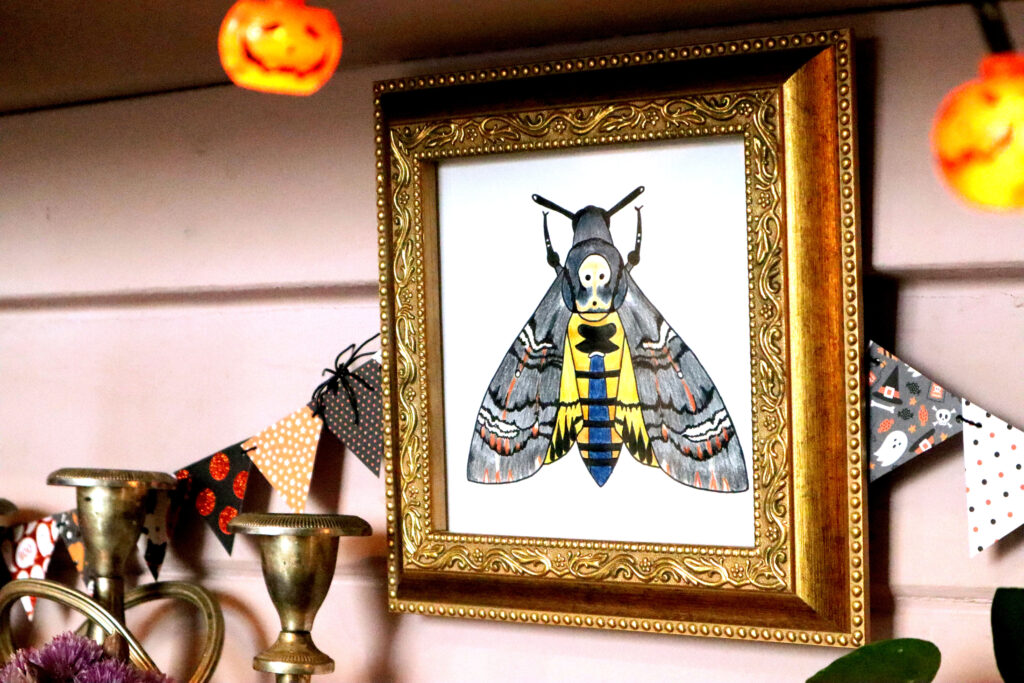 Image shows an original illustration of a death's head hawk moth in an ornate vintage gold frame. The frame hangs againt a pale pink wall. Behind the Fram hangs Halloween Mini Bunting. There are orange jack o'lantern fairy lights in the foreground. The top of a tarnished silver vintage candelabra is visible next to the frame.