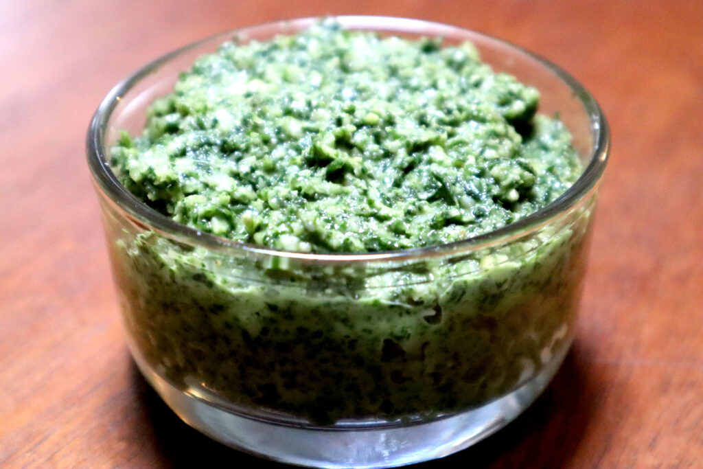 Image shows vibrant green fresh garden pesto in a glass dish. The dish sits on an antique timber table.