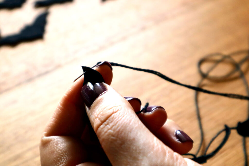 Closeup of Maddy the Maker's fingers holding a black felt bat. Maddy is wearing dark bergundy nailpolish. A sewing needle threaded with black cotton string has been pushed into the tip of the bat's second wing.