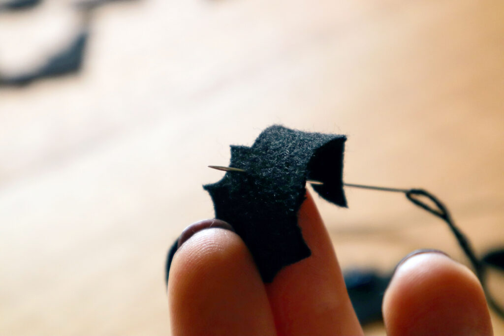 Closeup of Maddy the Maker's fingers holding a black felt bat. Maddy is wearing dark bergundy nailpolish. A sewing needle threaded with black cotton string has been pushed in throuh the tip of the bat wing, and out through the bat's eye.