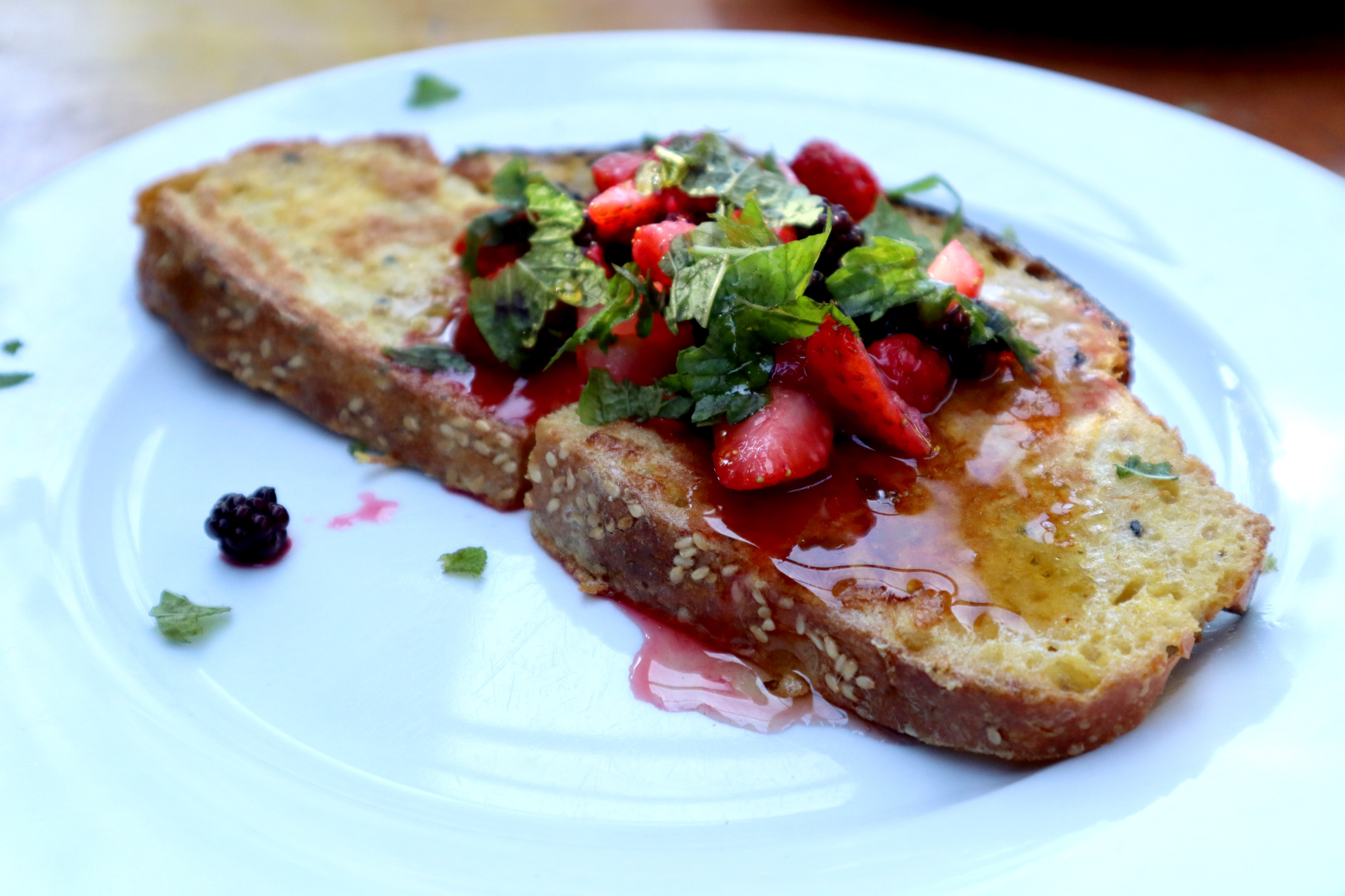 Image shows french toast on a white ceramic plate. The toast is topped with maple syrup, foraged blackberries, fresh home grown strawberries, raspberries, mint and lemon balm.