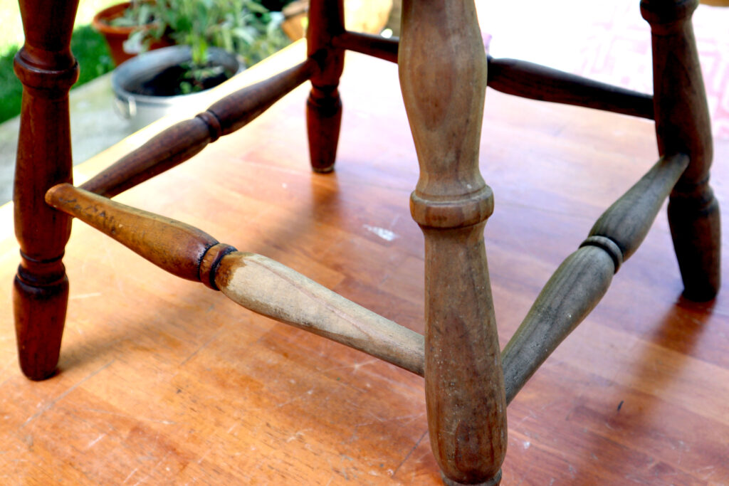 Detail of a vintage timber chair. Eco friendly wood polish has been applied to one part, here the wood looks clean and hydrated. Another section has not had the wood polish applied, here it looks dry and dirty. The chair is shown on a timber surface. In the background are some pots containing plants.