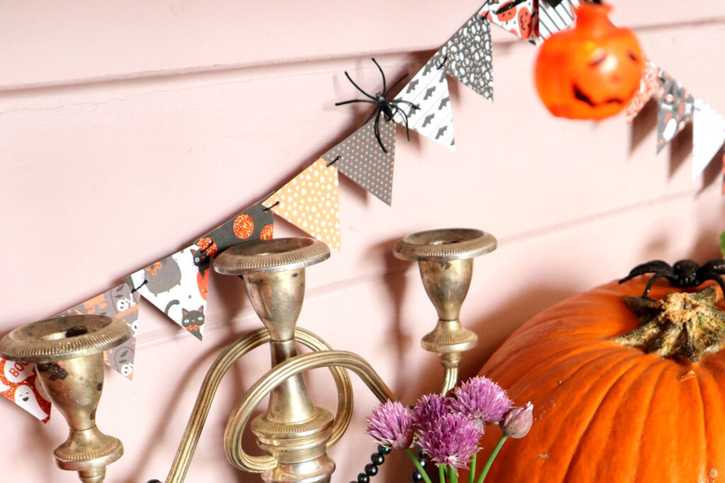 Closeup shot of Halloween Mini Bunting hanging against a pale pink wall. Each bunting triangle is made of patterned Halloween themed cardstock in a different design The coulours of the patterns are coordinating shades of orange, grey, black and white. There is a small black plastic spider hooked over the bunting. In front of the bunting, the tops of a vivid orange pumpkin and a tarnished vintage silver caldelabra are visible. There is a blak rubber spider on top of the pumpkin. In front of the candelabra, purple chive flowers are visible. At the top of the image, in the foreground, out of focus, hangs a vivid orange jack o'lantern fairy light.