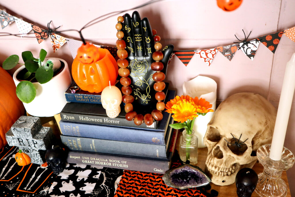 Image shows Halloween Mini Bunting hanging against a pale pink wall. There are two small black plastic spiders hooked over the bunting. In front of the wall is a halloween display. There is a black resin palmistry hand draped in a vintage orange bakelite necklace. The hand is next to a jack o'lantern candle, they sit atop a stack of fabric covered vintage books in indigo shades. To the left of the books is a potplant, a gravestone candle and an orange pumpkin. To the right of the books is a vintage glass inkwell containing vivid orange calendula flowers. Behind the vase is a white pilar candle. In front of the vase is a raw amethyst crystal. Next to the inkwell, is a realistic resin skull with a black plastic spider in its eye socket. In front of the skull is a small black skull candle, and a glass candlestick holding a tall white candle. A Halloween Table runner is also visible, it sits on a timber-look laminate surface.