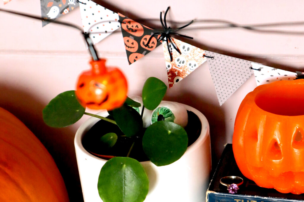 Closeup shot of Halloween Mini Bunting hanging against a pale pink wall. Each bunting triangle is made of patterned Halloween themed cardstock in a different design The coulours of the patterns are coordinating shades of orange, grey, black and white. There is a small black plastic spider hooked over the bunting. In front of the bunting is a small white planter pot containing a chinese money plant. Nestled behind the leaves are two hand painted air-dry clay eyeballs. To the left of the pot, part of an orange pumpkin is visible. To the right f the pot, an orange jack o'lantern candle is visible, next to it is a vintage amethyst ring. In the forground, out of focus, hangs an orange jack o'lantern fairy light.