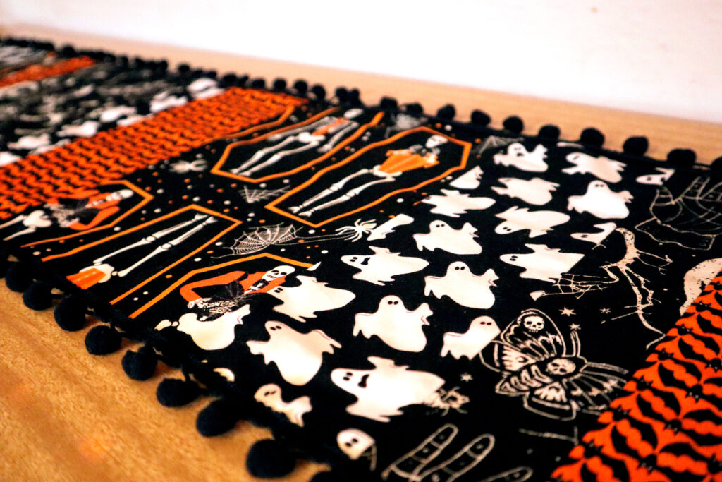 Closeup of Vintage halloween Table Runner. The table runner is made of strips of different halloween fabrics in coordinating black, white, and vivid orange. The edges of the table runner are decorated with black bobble fringe. Closeup of Vintage halloween Table Runner. The table runner is made of strips of different halloween fabrics in coordinating black, white, and vivid orange. The edges of the table runner are decorated with black bobble fringe. The table runner is laid out on top of a 60s laminate buffet top.