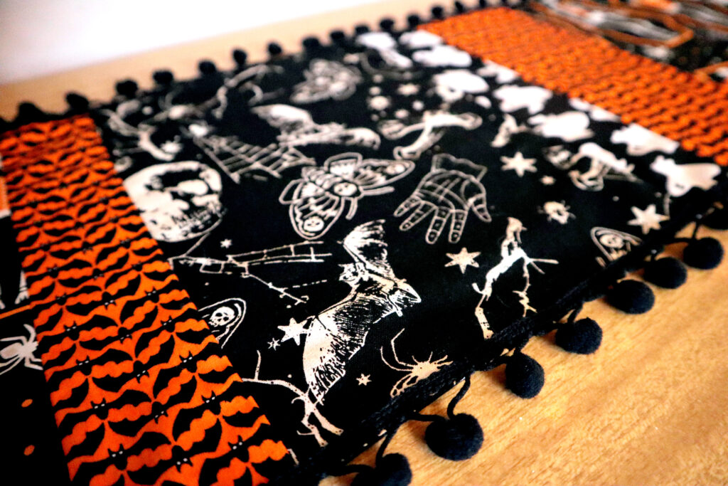 Closeup of Vintage halloween Table Runner. The table runner is made of strips of different halloween fabrics in coordinating black, white, and vivid orange. The edges of the table runner are decorated with black bobble fringe. The table runner is laid out on top of a 60s laminate buffet top.