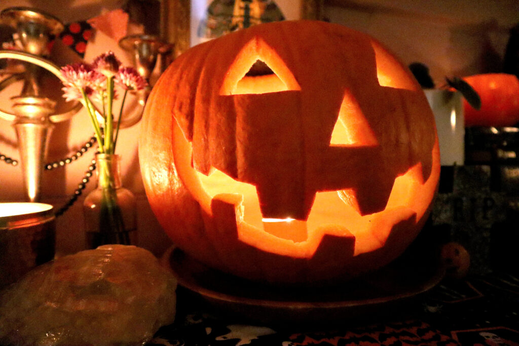 Image shows a large, smiling jack o'lantern in a dimly lit room. The candle inside is lit and the toothy grin of the jack o'lantern glows. To the left of the jack o'lantern a small vintage glass bottle is visible. The bottle contains purple chive flowers. Next to the bottle is a vintage silver candelabra with a strand of black pearls hanging from it. In front of the candelabra, a lit candle in a tin is visible.