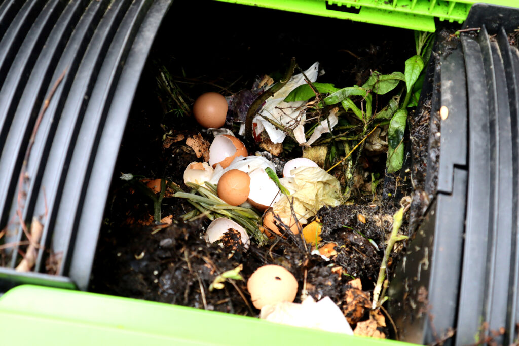 Image shows an open rotating black compost bin. Inside the compost bin is black compost, eggshells, paper, green plant material, and other assorted food scraps. 