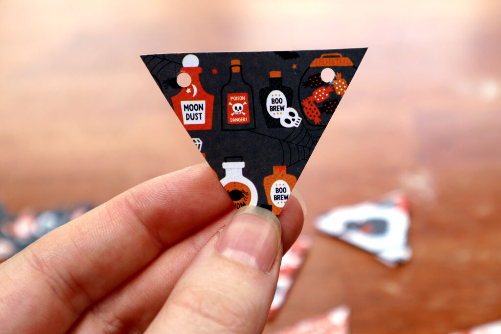 Image shows Maddy the Maker's fingertips holding an inverted triangle of cut out halloween cardstock by the bottom point. There is a hole punched in each of the top points of the triangle. in the background, out of focus, a timber surface with other cut paper triangles on it is visible.