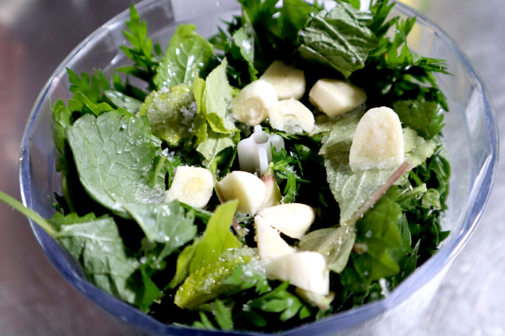 Image shows a small food processor filled with fresh green leafy herbs, sliced garlic, olive oil, and salt.