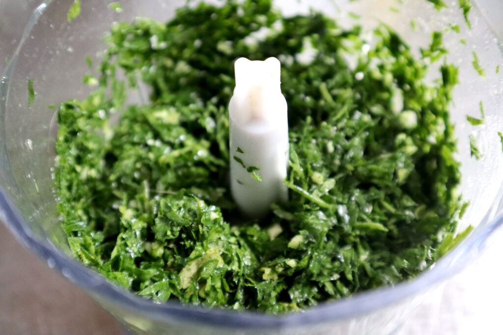 Image shows a small food processor filled with coarsely chopped fresh herbs. 