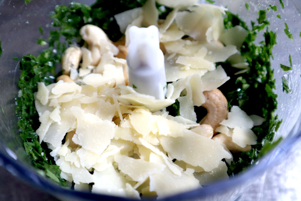 Image shows a small food processor filled with chopped herbs, cashew nuts and shaved parmesan cheese.