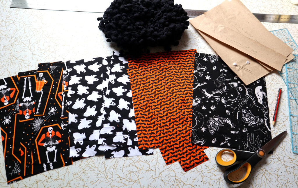 Items tequired to make a Vintage Halloween Table Runner are arranged on a retro white laminate tabletop with gold and black patterning. There are paper pattern pieces, pieces of halloween fabrics in four different prints, a metal dressmakers ruler, clear plastic quilting rulers, a ball of black bobble fringe, Black and orange fabric scissors, a greylead pencil, and two pins with white plastic hearts at the top.