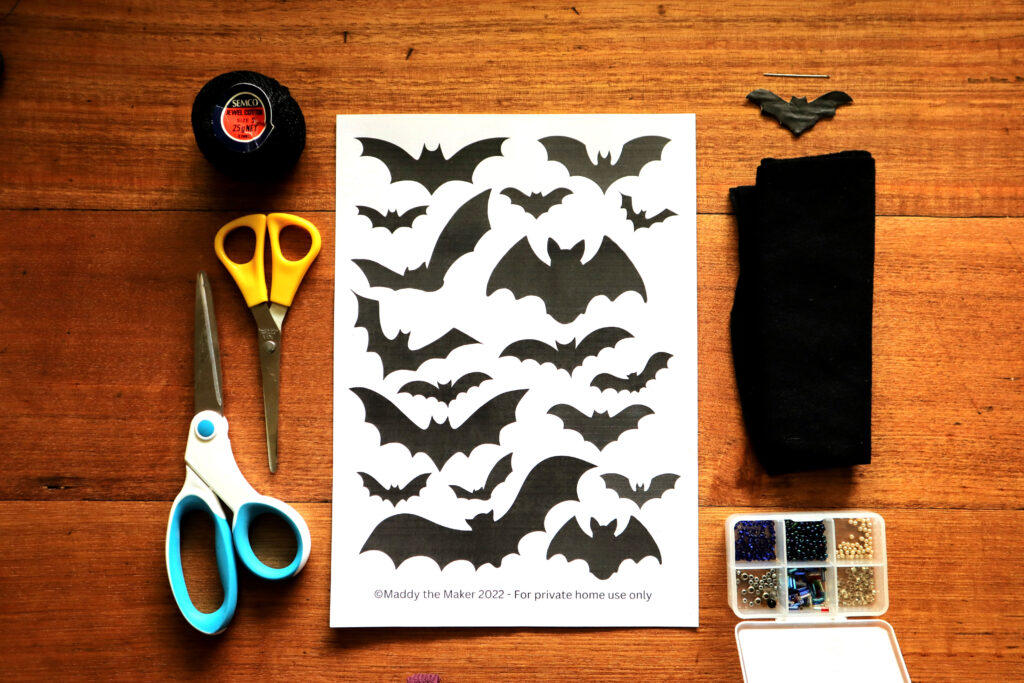 Image shows the materials required to make a Halloween Felt Bat Garland. These are Maddy the Maker's free bat template printable, a ball of thrifted black cotton string, a yellow pair of scissors for cutting paper, a white and blue pair of scissors for cutting felt, an large sewing needle, a cardboard bat template, a piece of black wool felt, and a small plastic box containing iridescent beads. The items are shown from above, they are lying on a vintage timber desk.