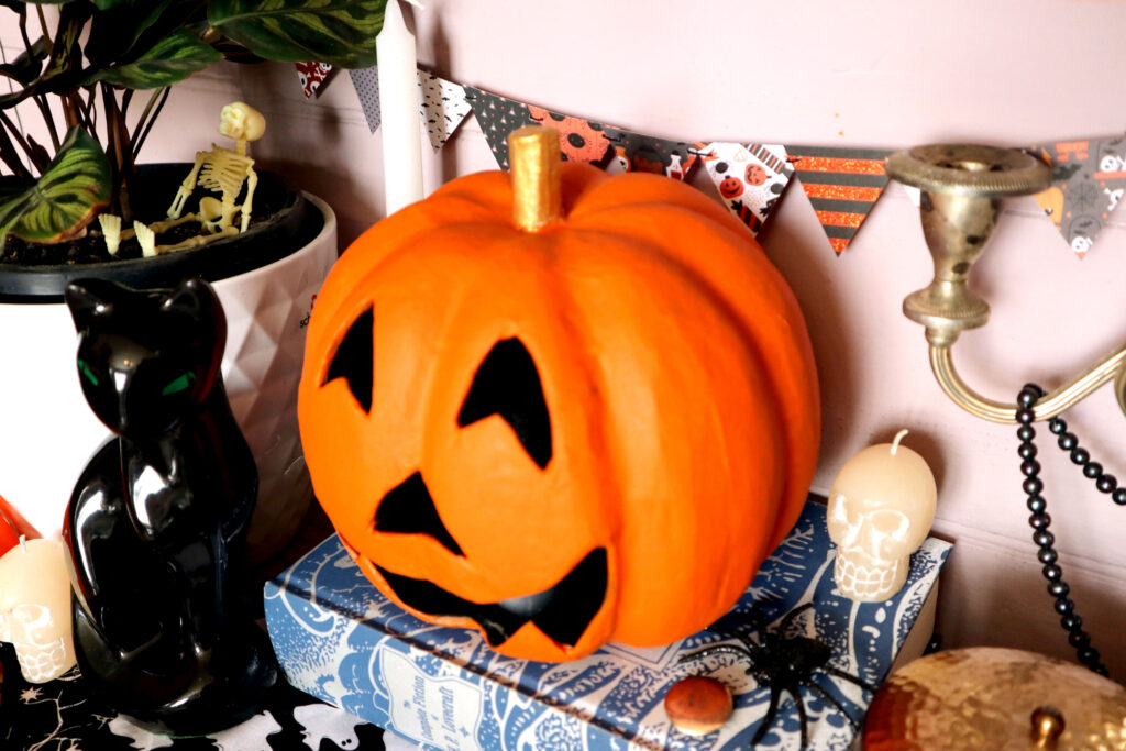 Image shows a vivid orange hand painted papier mache jack o'lantern,, with a gold stem. The jack o'lantern sits on a blue and silver fabric covered edition of H.P. Lovecraft's collected works. On the book next to the jack o'lantern is a small bone coloured skull candle, a glittery rubber spider, and a vintage monogrammed copper brooch. On the left of the jack o'lantern is a prayer plant in a white ceramic pot. There is a small plastic skeleton sitting in the pot. In front of the pot is a vintage black ceramic cat figurine and another skull candle. To the right of the jack o'lantern part of a tarnished silver vintage candelabra is visible. There is a strand of black pearls hanging from the candelabra. The top of a round beaten-brass tin is also visible. On the light pink wall behind the jack o'lantern hangs a strand of Halloween Mini Bunting.