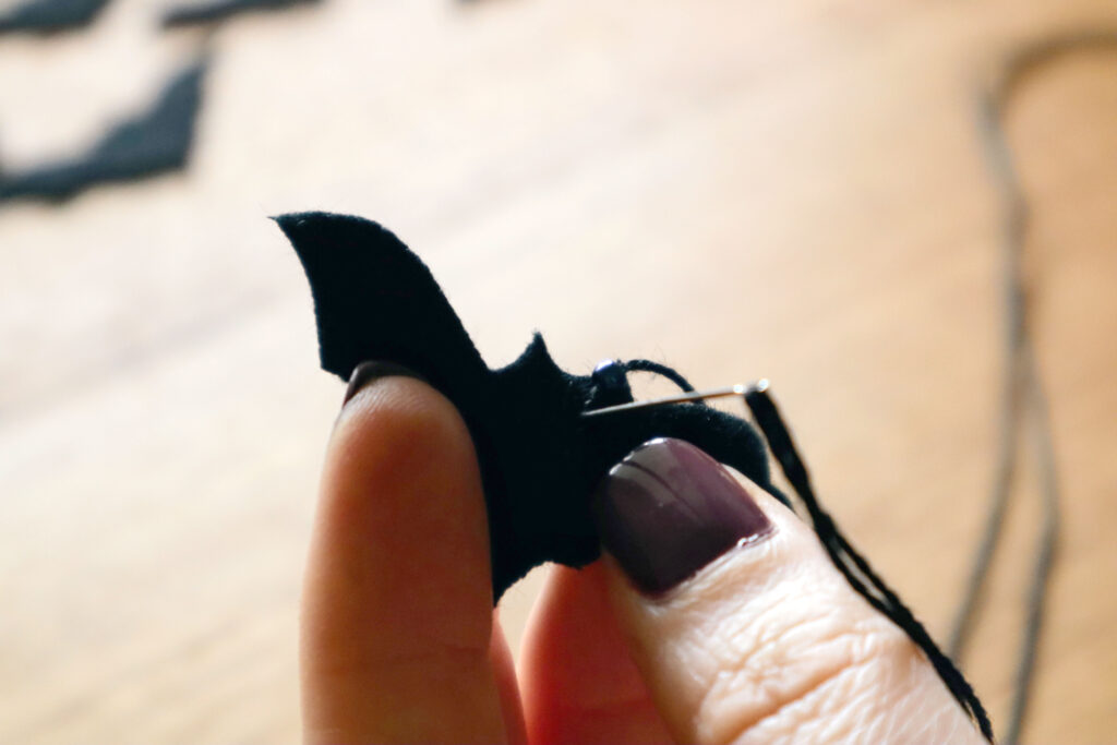 Closeup of Maddy the Maker's fingers holding a black felt bat. Maddy is wearing dark bergundy nailpolish. A sewing needle threaded with black cotton string has been pushed out through the bat's eye. An iridescent bead has been threaded onto the string, before pushing the needle back through the bat's eye.