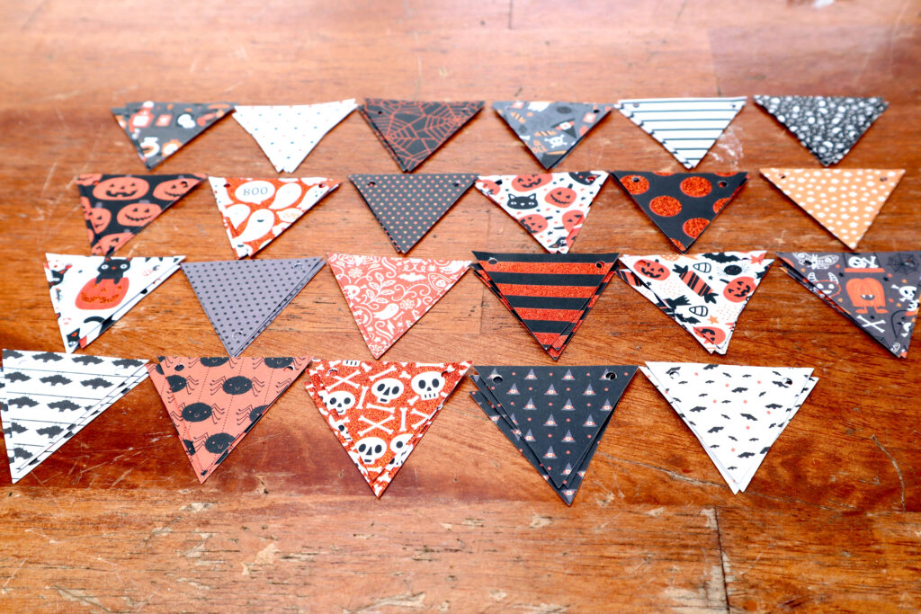 Image shows a scratched timber surface covered in small inverted triangles cut from Halloween cardstock. The triangles are arranged into small piles, with each design having its own pile. There are 23 different designs. They are arranged in four neat rows, with the upper corners touching.