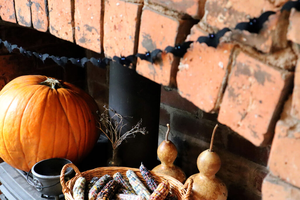 Sustainable Batty halloween Garland is shown hanging against a rustic brick chimney. The bats are made of upcycled black wool felt. Their eyes are shiny irridescent glass beads which reflect the light. The bats are strung on thrifted black cotton cord. Inside the chimney sits a black cast iron wood-burning stove. On top of the stove is a large, bright orange pumpkin, a small vintage glass bottle with dried raspberry prunings, two dried bottle gourds, a basket of rainbow coloured glass gem corn, and a small cast iron cauldron.