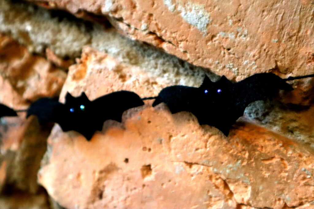 Closeup shot of DIY Halloween felt bat garland, shown hanging against a rustic brick chimney. The bats are made of upcycled black wool felt. Their eyes are shiny irridescent glass beads which reflect the light. The bats are strung on thrifted black cotton cord.