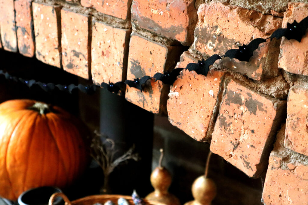 Sustainable Batty Halloween Garland is shown hanging against a rustic brick chimney. The bats are made of upcycled black wool felt. Their eyes are shiny irridescent glass beads which reflect the light. The bats are strung on thrifted black cotton cord. Inside the chimney sits a black cast iron wood-burning stove. On top of the stove is a large, bright orange pumpkin, a small vintage glass bottle with dried raspberry prunings, two dried bottle gourds, a basket of rainbow coloured glass gem corn, and a small cast iron cauldron.