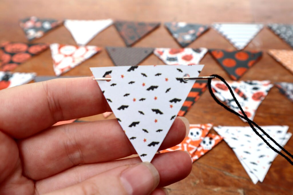 Image shows Maddy the Maker's hand holding a Halloween cardstock inverted triangle with a hole in each top corner. There is a large needle threaded with black cotton string which has been pushed through one hole and out the other. In the background a timber surface covered in rows of Halloween cardstock inverted triangles is visible.