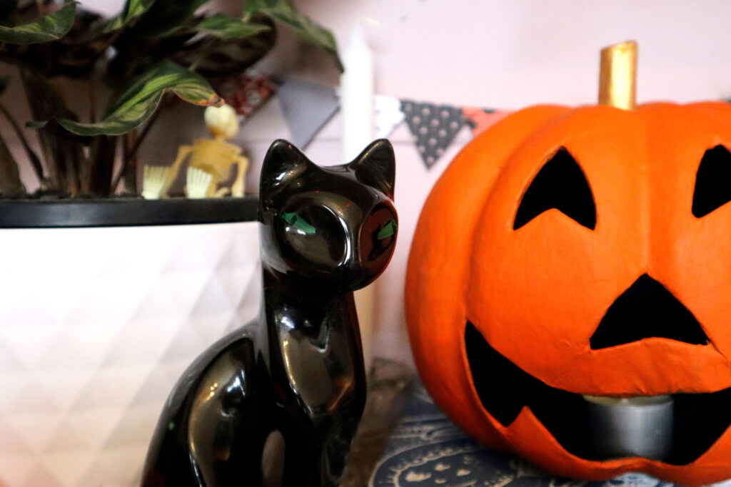 Image shows a closeup of a vintage black ceramic cat figurine with green eyes. Behind the cat is a white taper candle and a ceramic plant pot containing a prayer plant and a small plastic skeleton. To the right of the cat, part of a vivid orange hand-painted papier mache jack o'lantern is visible. On the pale pink wall behind the cat hangs a string of Mini Halloween Bunting.