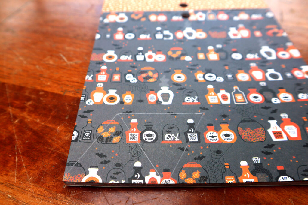 Image shows a pad of halloween themed cardstock, lying on a timber surface. The paper shown is charcoal coloured. The repeating pattern shows various jars full of witchy ingredients candy, and bones. There are also spiderwebs and bats. At the bottom of the paper, there is a tesselating pattern of shapes drawn in greylead pencil. In the bottom left corner is a pyramid shaped triangle. There is an inverted triangle right next to it.