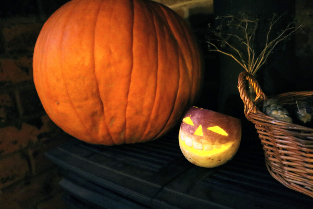 Image shows a dimly lit scene containing a large orange pumpkin and a small carved turnip tumshie head. The tumshie contains a lit candle, making its features glow yellow. Nexy to the tumshie is a wicker basket ontaining glass gem corn. Behind the basket is a small vintage glass bottle containing dead twigs.