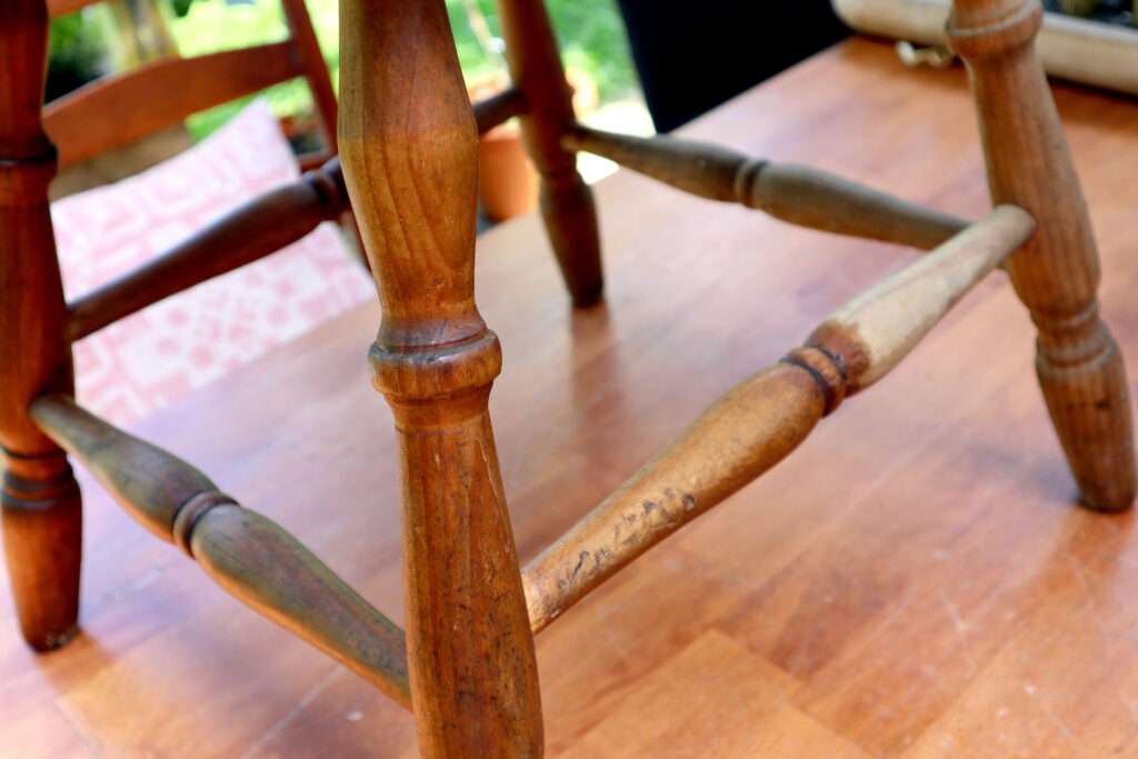 Detail of a vintage timber chair. Eco friendly wood polish has been applied to one part, here the wood looks clean and hydrated. Another section has not had the wood polish applied, here it looks dry and dirty. The chair is shown on a timber surface. In the background are some pots containing plants.