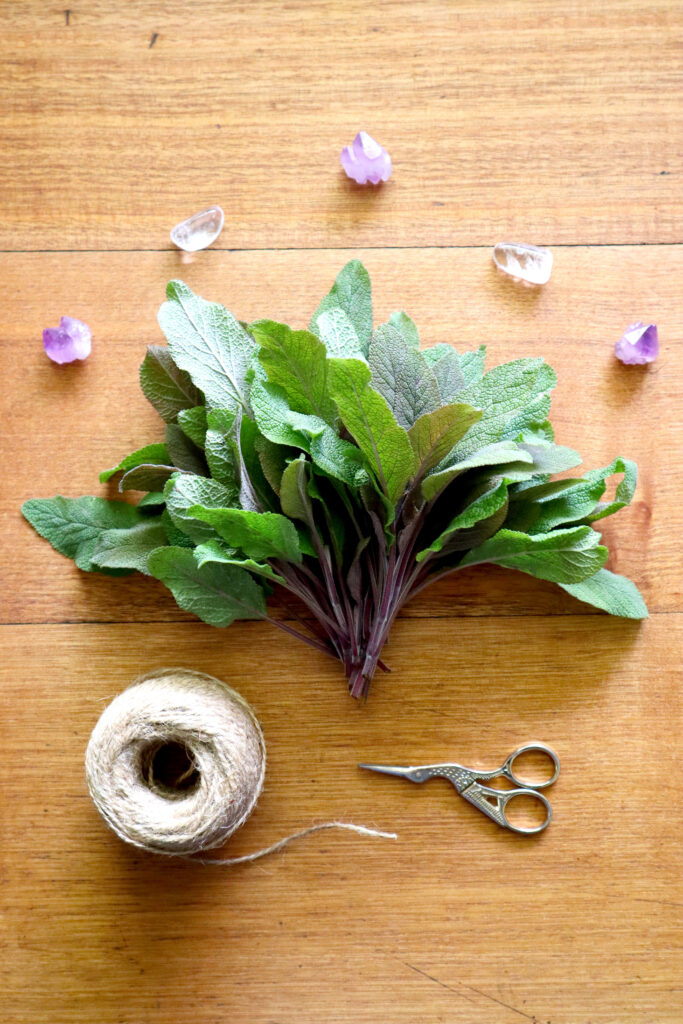 Image shows a vintage timber desk from above. On the desk is a bunch of freshly harvsted purple sage. There is also a ball of jute twine and a small pair of gold scissors. Above the bunch of sage, there are three raw amethyst crystals and two polished clear quartz crystals.