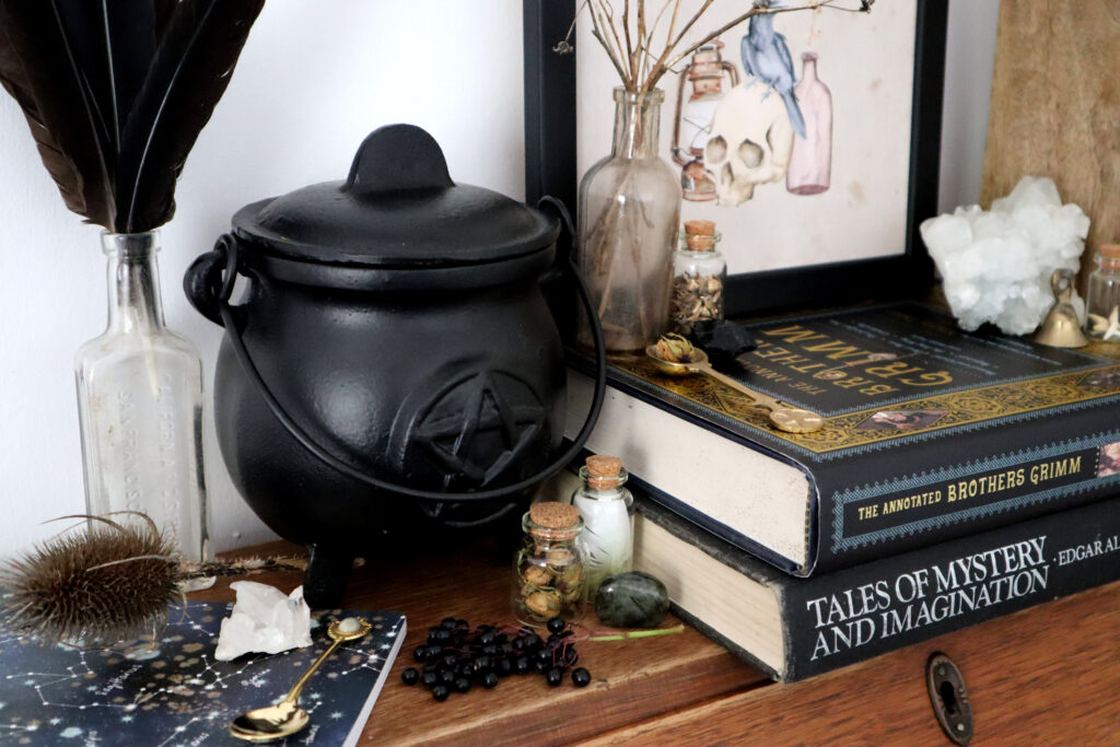 Image shows a witches altar. Items shown are a black cauldron with lid, vintage medicine bottle with black raven feathers, assorted small glass jars with cork stoppers containing items collected from nature, raw crystals, vintage Poe and Grimm hardback books, a framed print, raw and tumbled crystals, a vintage gold agate spoon teaspoon, a brass teaspoon, a teasel comb, and a bunch of fresh elderberries.