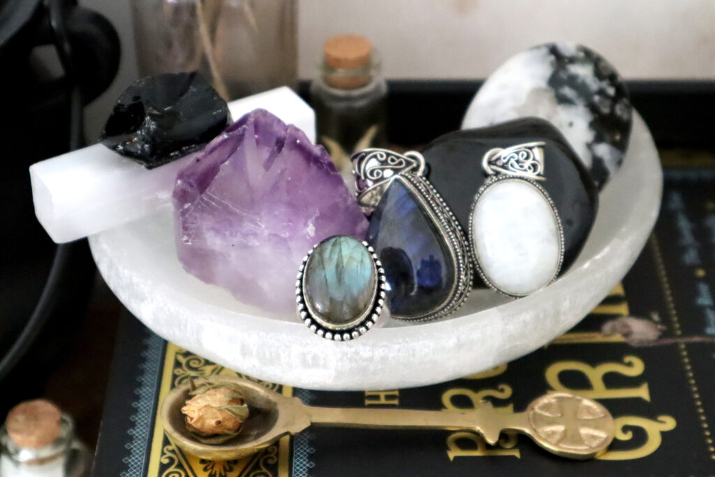 Image shows a white selenite moon shaped dish on a vintage book. The dish contains other crystals and jewellery. These include a stick of white satin spar, raw obsidian, raw amethyst, a labradorite ring and pendant, a moonstone pendant, polished black tourmaline, and a rainbow moonstone palm stone with black tourmaline inclusions. In front of the moon dish is a small brass teaspoon containing a dried rosebud.