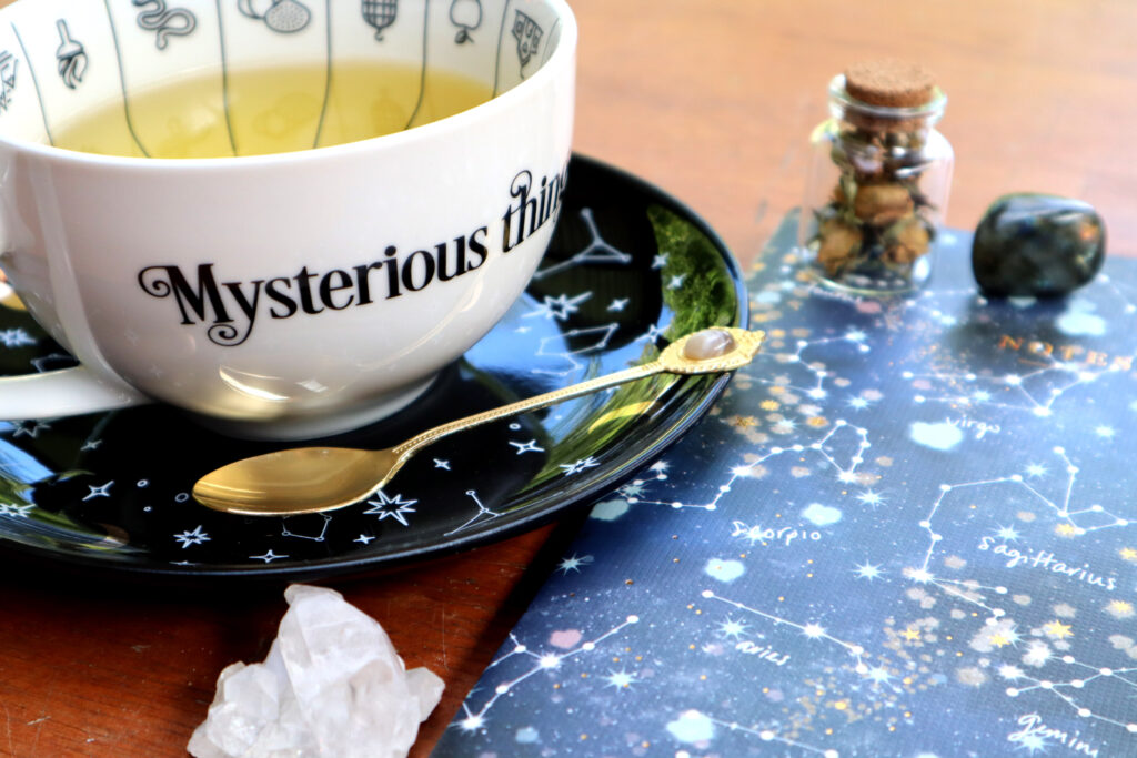 Image shows items used for a Healing Tea Ritual. These include a  witchy teacup full of herbal tea with matching saucer, a vintage gold agate teaspoon, a small jar of dried rose buds with a cork stopper, a labradorite tumbled crystal, and a raw himalayan quartz crystal.