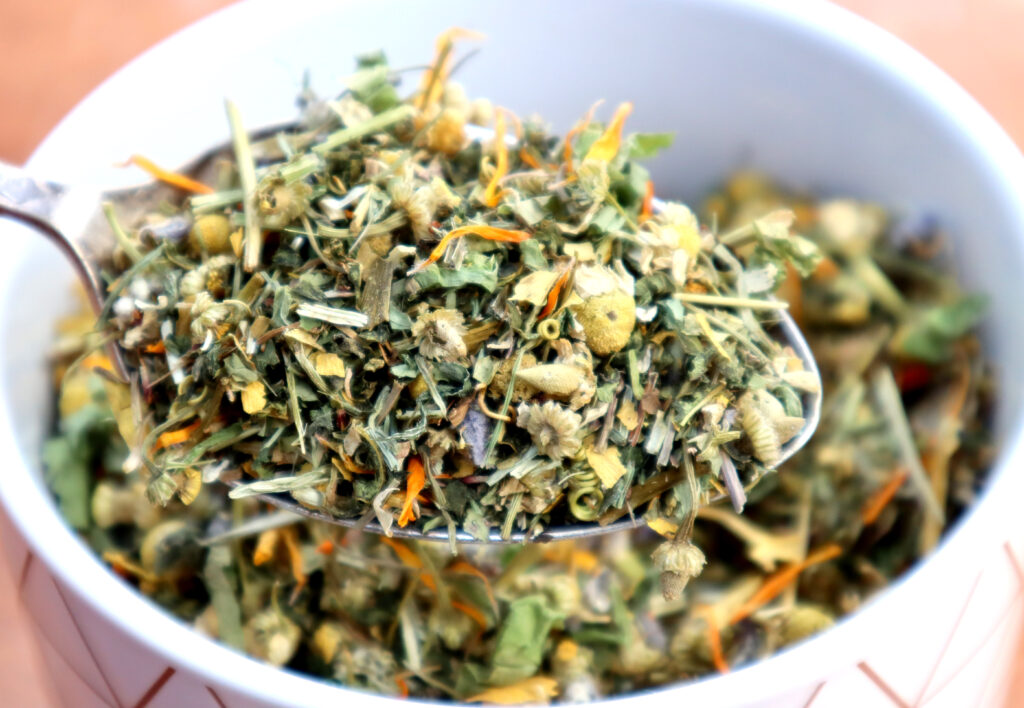 Nervous/Immune System Healing Tea is shown in a white ceramic canister with copper geometric markings. Loose tea is shown close up on a vintage silver spoon. The spoon is held above the ceramic canister for a better view. The dried herbs included are shades of green, brown, yellow, orange and purple.