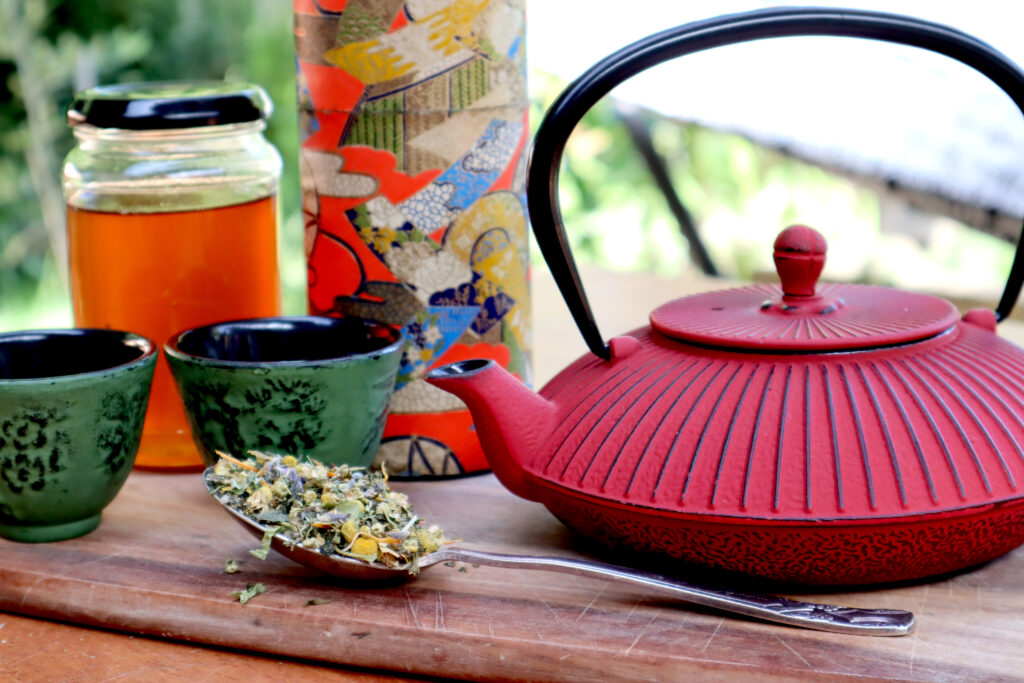 Nervous and Immune System Balancing Tea is shown outdoors on a rustic timber surface, with greenery in the background. Loose tea is shown on a vintage silver spoon. There is also a red cast iron teapot, two green cast iron cups, a vintage japanese tea canister in several colours, and a clear glass jar of honey.