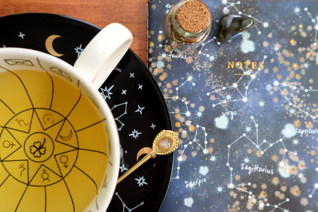 Closeup shot taken from above. There is a witchy white tasseomancy teacup containing herbal tea, a matching black saucer with stars and constellations, a vintage gold agate spoon, a blue zodiac notebook, small jar containing dried rosebuds with cork stopper, and a tumbled labradorite crystal.